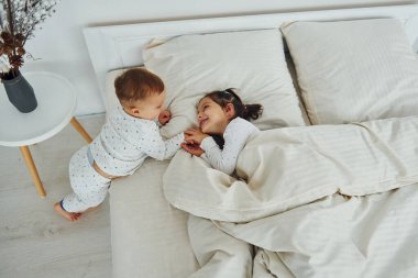 Two kids have fun while liyng down on bed. Interior and design of beautiful modern bedroom at daytime.