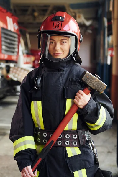 With hammer in hands. Female firefighter in protective uniform standing near truck.