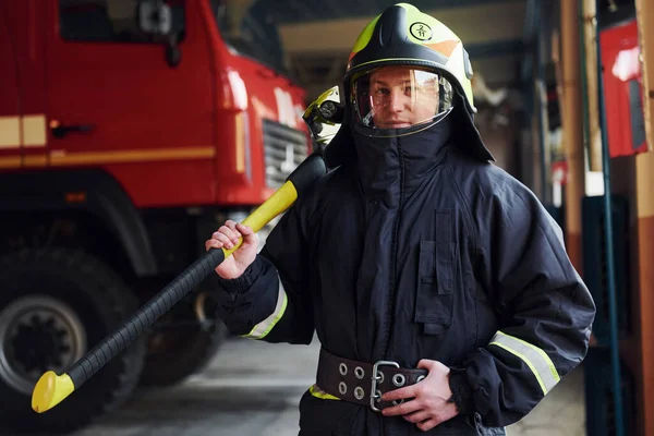 Holds axe in hands. Male firefighter in protective uniform standing near truck.