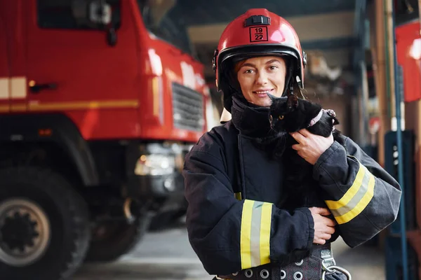 Cute black cat. Female firefighter in protective uniform standing near truck.