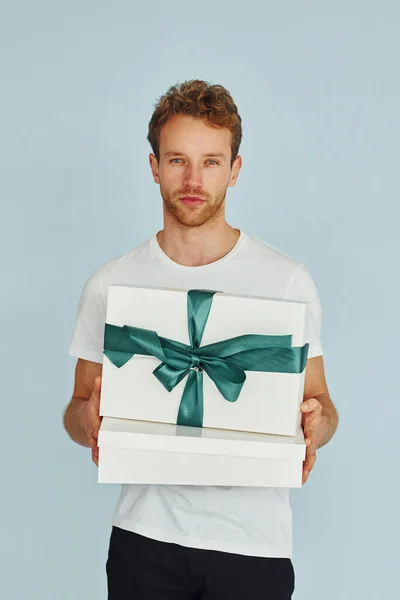 Young man in white shirt standing against wall and holds gift box.