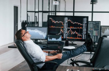 Sandalyeye oturur. Genç borsacı erkek ofis içinde çalışıyor. Ekranların çoğu.