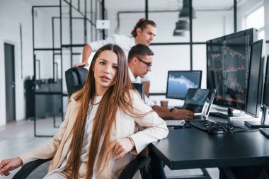 Kadın kameraya bakıyor. Borsacılardan oluşan bir ekip modern ofiste birçok ekran ekranı ile çalışır..