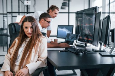Kadın kameraya bakıyor. Borsacılardan oluşan bir ekip modern ofiste birçok ekran ekranı ile çalışır..