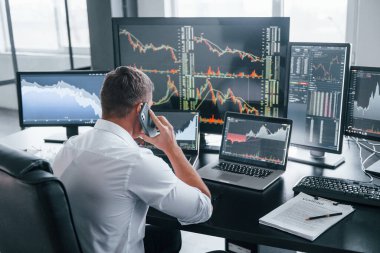 Telefonla konuşuyor. Genç borsacı erkek ofis içinde çalışıyor. Ekranların çoğu.