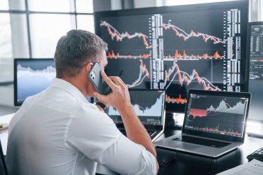 Telefonla konuşuyor. Genç borsacı erkek ofis içinde çalışıyor. Ekranların çoğu.