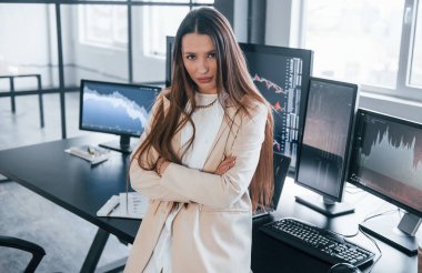Kameraya poz ver. Genç bayan borsacı ofisin içinde. Ekranların çoğu.