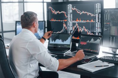 Resmi kıyafetlerle. Genç borsacı erkek ofis içinde çalışıyor. Ekranların çoğu.