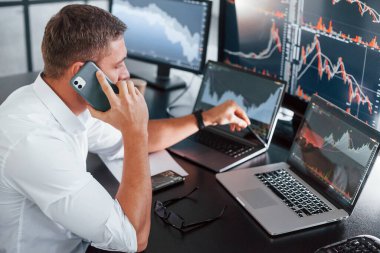 Telefonla konuşuyor. Genç borsacı erkek ofis içinde çalışıyor. Ekranların çoğu.
