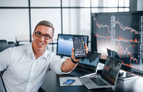 Telefonu tutuyor. Genç borsacı erkek ofis içinde çalışıyor. Ekranların çoğu.