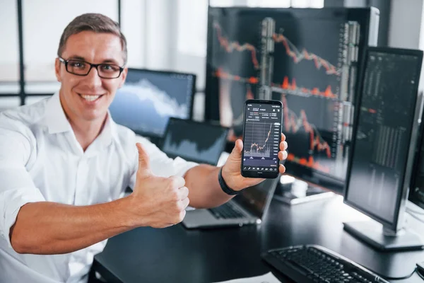 Telefonu tutuyor. Genç borsacı erkek ofis içinde çalışıyor. Ekranların çoğu.