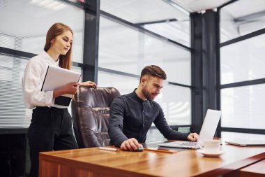 Resmi giyinmiş bir kadın ve bir adam ofis içinde masa başında birlikte çalışıyorlar..