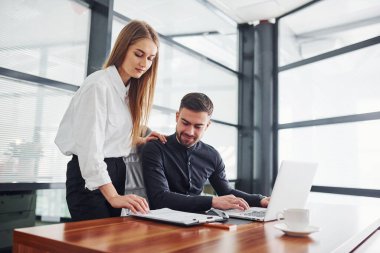 Resmi giyinmiş bir kadın ve bir adam ofis içinde masa başında birlikte çalışıyorlar..