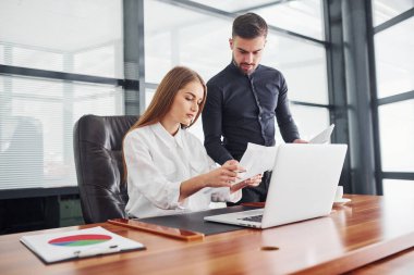 Resmi giyinmiş bir kadın ve bir adam ofis içinde masa başında birlikte çalışıyorlar..