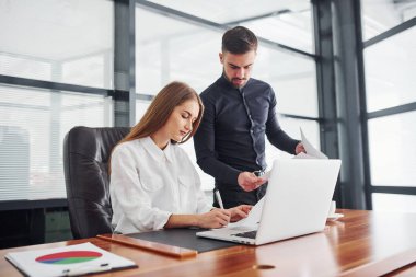 Resmi giyinmiş bir kadın ve bir adam ofis içinde masa başında birlikte çalışıyorlar..