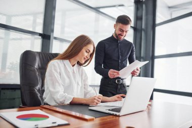 Resmi giyinmiş bir kadın ve bir adam ofis içinde masa başında birlikte çalışıyorlar..