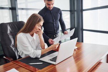 Resmi giyinmiş bir kadın ve bir adam ofis içinde masa başında birlikte çalışıyorlar..