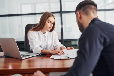 Belgelerle oturan adam. Resmi giyinmiş bir kadın ve bir adam ofis içinde masa başında birlikte çalışıyorlar..
