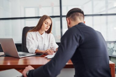 Belgelerle oturan adam. Resmi giyinmiş bir kadın ve bir adam ofis içinde masa başında birlikte çalışıyorlar..