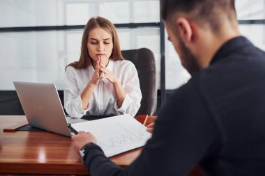 Belgelerle oturan adam. Resmi giyinmiş bir kadın ve bir adam ofis içinde masa başında birlikte çalışıyorlar..