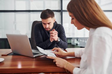 Resmi giyinmiş bir kadın ve bir adam ofis içinde masa masa dokümanlarla birlikte çalışıyorlar..