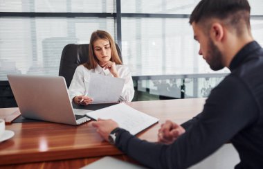 Belgelerle oturan adam. Resmi giyinmiş bir kadın ve bir adam ofis içinde masa başında birlikte çalışıyorlar..