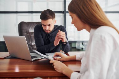 Resmi giyinmiş bir kadın ve bir adam ofis içinde masa masa dokümanlarla birlikte çalışıyorlar..