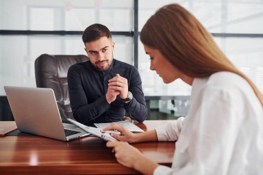 Resmi giyinmiş bir kadın ve bir adam ofis içinde masa masa dokümanlarla birlikte çalışıyorlar..