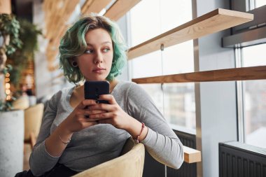 Alternatif kız, yeşil saçlı, gündüz vakti evde oturan, elinde telefon olan..