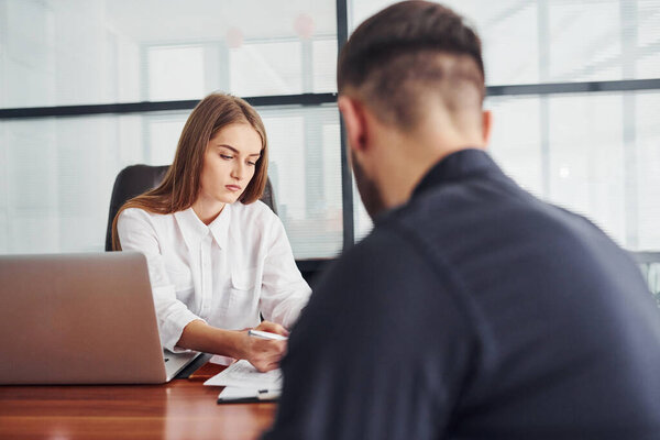 Guy sitting with documents. Woman and man in formal clothes working together indoors in the office by table.