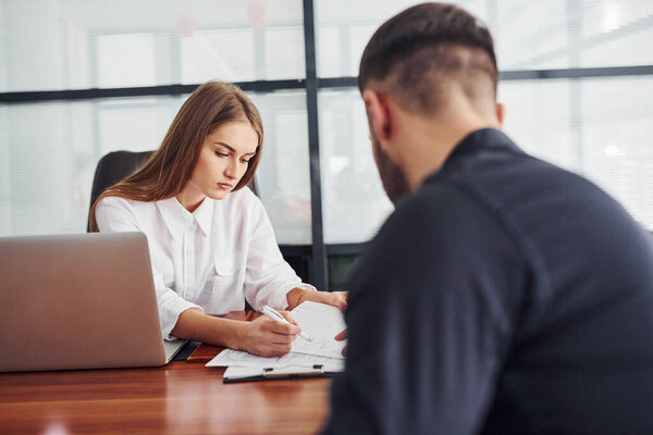 Guy sitting with documents. Woman and man in formal clothes working together indoors in the office by table.