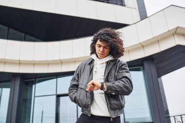 Handsome young man with curly black hair posing for the camera on the street against building.