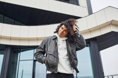 Kıvırcık siyah saçlı yakışıklı genç adam sokakta binaya karşı kameraya poz veriyor..