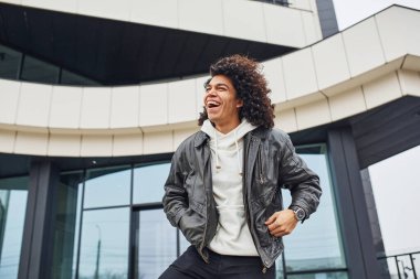 Kıvırcık siyah saçlı yakışıklı genç adam sokakta binaya karşı kameraya poz veriyor..