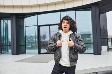 Kıvırcık siyah saçlı yakışıklı genç adam sokakta binaya karşı kameraya poz veriyor..