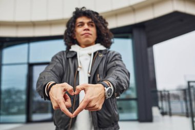 Kıvırcık siyah saçlı yakışıklı genç adam sokakta binaya karşı kameraya poz veriyor..