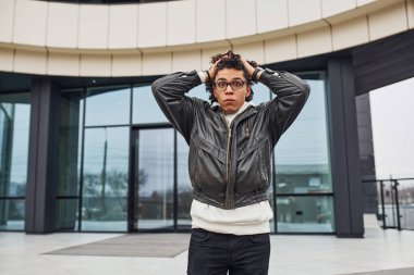 Kıvırcık siyah saçlı yakışıklı genç adam ayakta duruyor ve caddede bina karşısında şoka uğramış hissediyor..