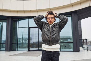 Kıvırcık siyah saçlı yakışıklı genç adam ayakta duruyor ve caddede bina karşısında şoka uğramış hissediyor..