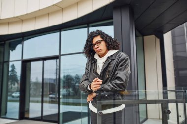 Kıvırcık siyah saçlı ve gözlüklü yakışıklı genç adam caddede binaya karşı..