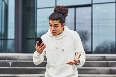 Kıvırcık siyah saçlı yakışıklı genç adam caddede binanın karşısında telefonla konuşuyor..