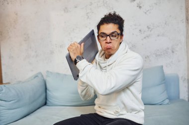 Kıvırcık siyah saçlı kızgın genç adam elinde dizüstü bilgisayarla çılgına dönüyor ve sinirleniyor..