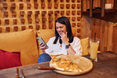 Genç esmer kız elinde pizza ve akıllı telefonuyla evde oturuyor..