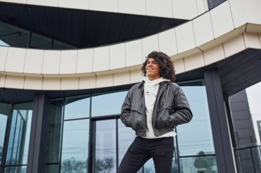 Kıvırcık siyah saçlı yakışıklı genç adam sokakta binaya karşı kameraya poz veriyor..