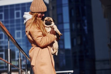 Sıcak giysili bir kadın, arka planda iş yeri yakınlarında köpeğini elinde tutuyor..