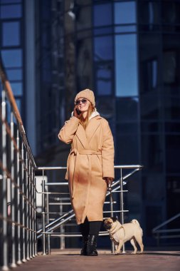 Sıcak giysili bir kadın iş yerinin yakınında köpeğiyle yürüyüşe çıkıyor. Arka planda bunu inşa ediyor ve telefonla konuşuyor..