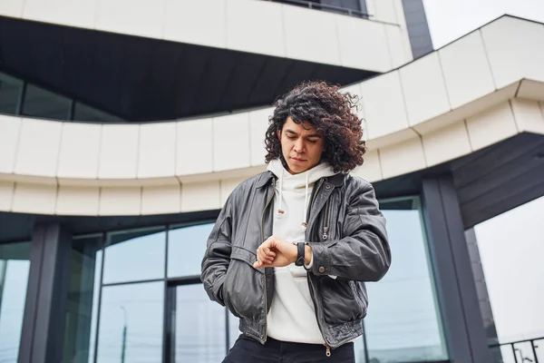 Kıvırcık siyah saçlı yakışıklı genç adam sokakta binaya karşı kameraya poz veriyor..