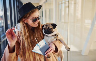 Young female passenger in warm clothes holding tickets and cute dog in hands in airport hall.