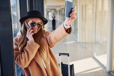Güneş gözlüklü, kalın elbiseli ve şapkalı genç bir kadın havaalanında elinde telefon ve biletlerle bekliyor..
