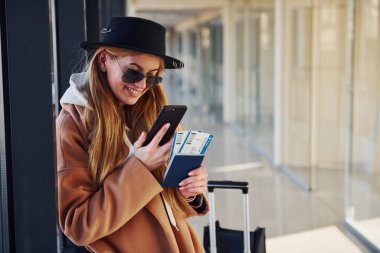 Güneş gözlüklü, kalın elbiseli ve şapkalı genç bir kadın havaalanında elinde telefon ve biletlerle bekliyor..