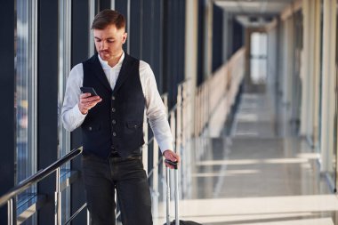 Young male passenger in elegant formal clothes is in the airport hall standing with baggage and phone in hand.